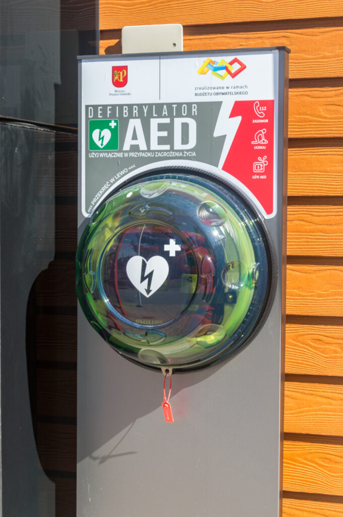 Defibrylator AED zamontowany na ścianie zewnętrznej budynku. Urządzenie oznaczone symbolem serca i pioruna, gotowe do użycia w sytuacjach nagłego zatrzymania krążenia.